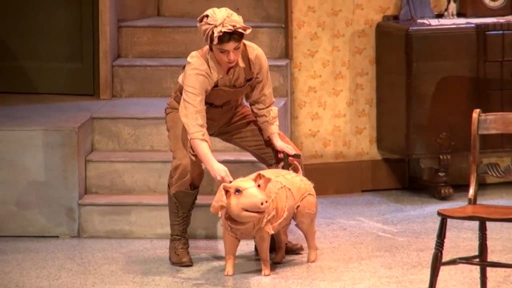 A woman in vintage attire kneels beside a crafted pig with big eyes on a stage