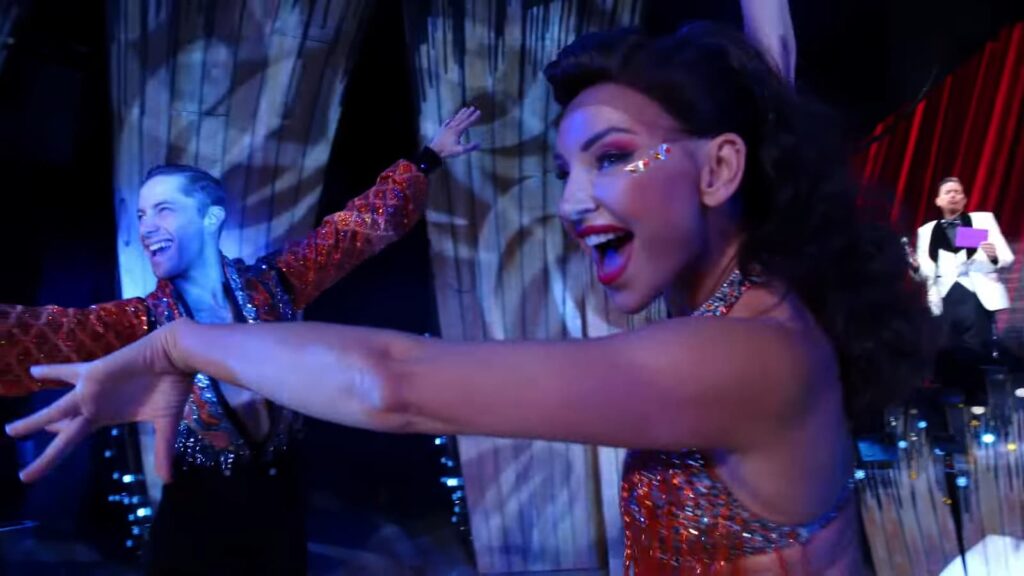 Joyful female dancer in red sparkles poses, with a male dancer and musician in the background