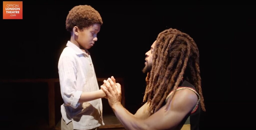 An artist dressed as Bob Marley plays a role with a boy on stage