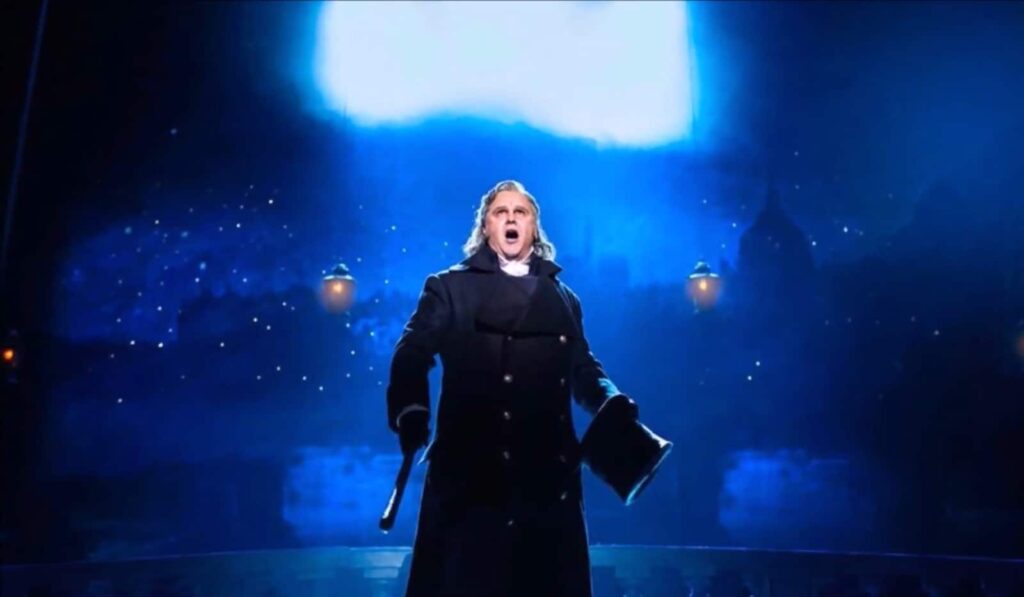 man in a black coat sings and holds a hat in his hand, the blue blurred image of city behind