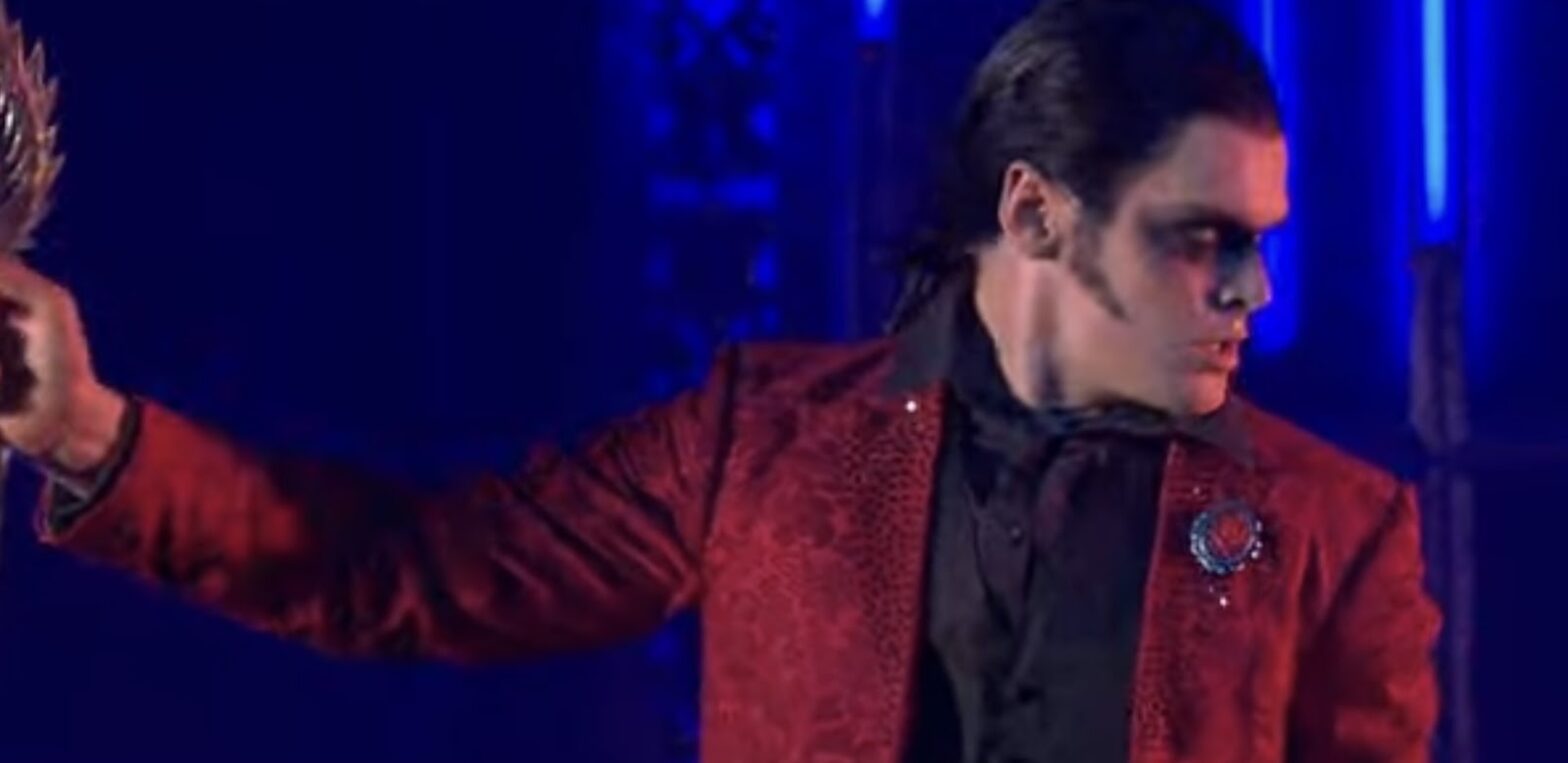 A man with face paint, wearing a red jacket and black shirt, holding an object